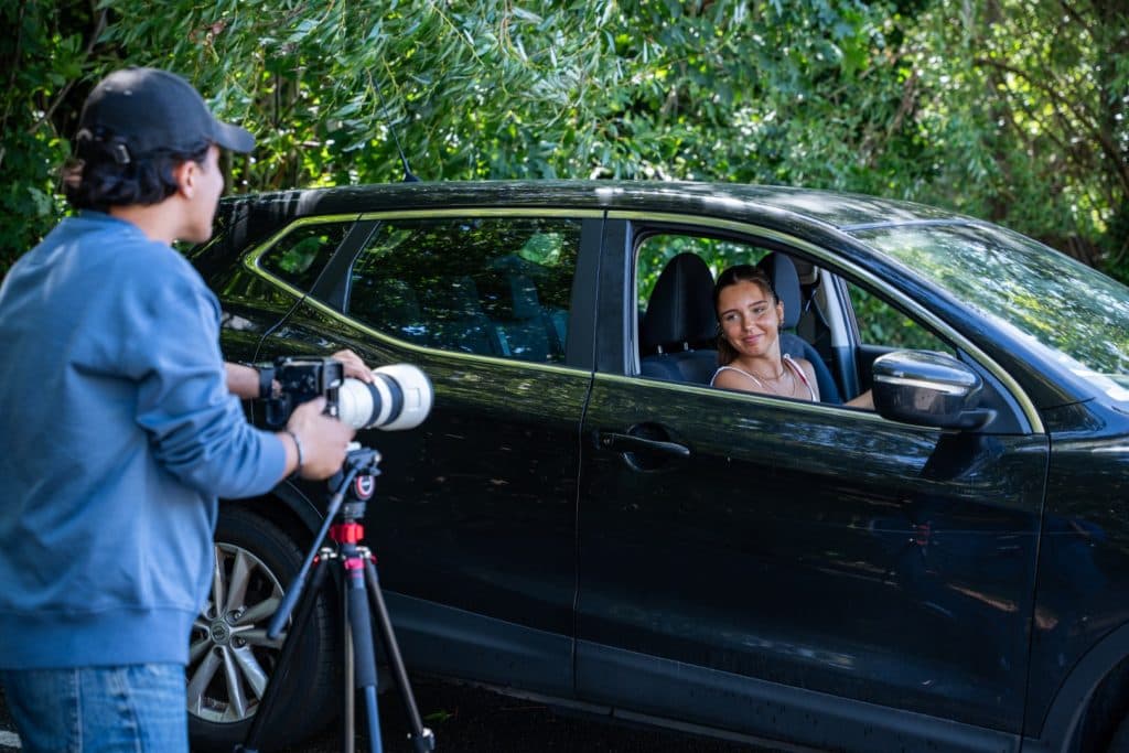Videographer filming a smiling woman sitting in a black car with a camera on a tripod during an outdoor shoot